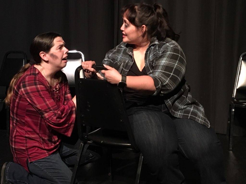 Dominica kneels on the floor with a scar on her forehead and is clearly distressed, while Alissa sits on a backwards chair and tries to comfort her.