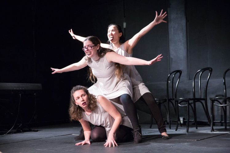 Casey Busher crouches on the floor while Dominica May and Shirley Chan spread their arms like wings above her